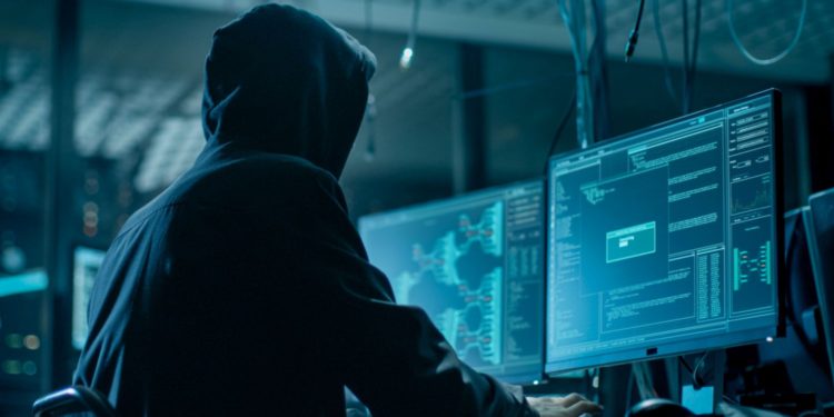 Shot from the Back to Hooded Hacker Breaking into Corporate Data Servers from His Underground Hideout. Place Has Dark Atmosphere, Multiple Displays, Cables Everywhere.